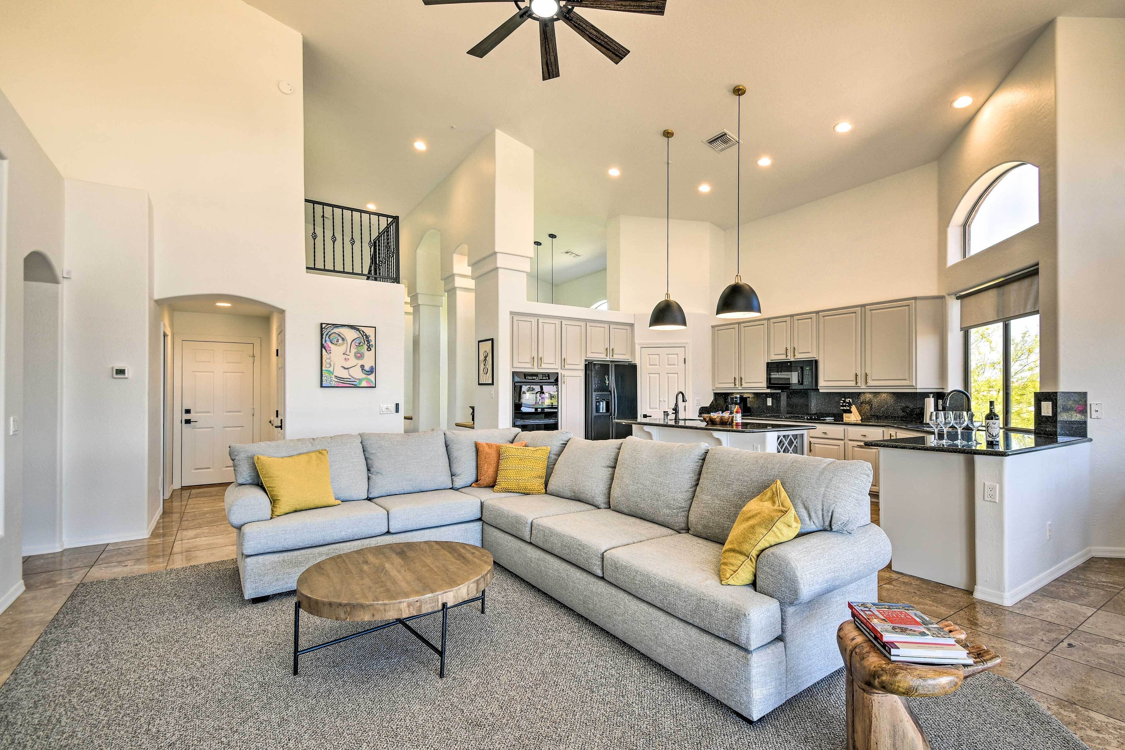 A spacious living room with a grey sectional sofa, a wooden coffee table, and a kitchen area with white cabinets and a black refrigerator.