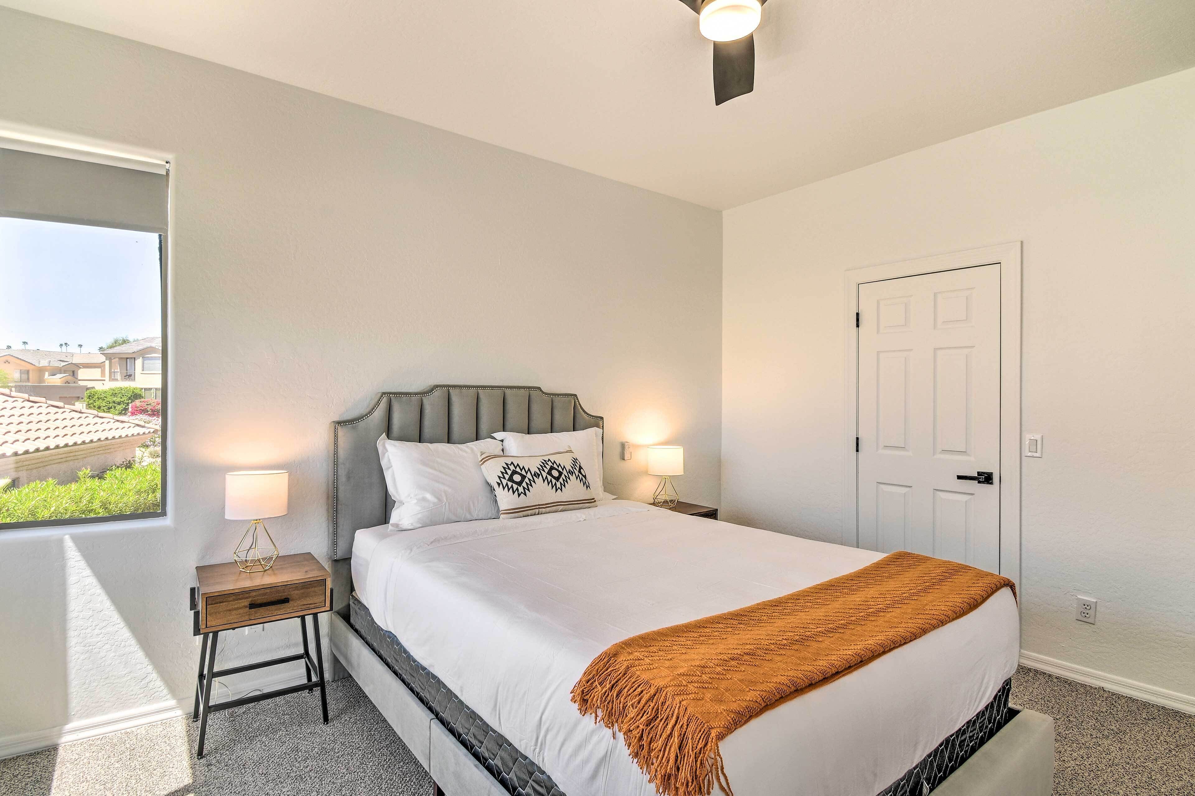A bedroom with a bed, bedside lamps, a nightstand, a door, and a window with a view of buildings.