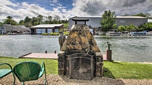 Une cheminée en pierre avec une porte en métal et un motif en étoile, installée sur une surface de gravier, avec une chaise verte et une jetée qui s’avance dans un lac.