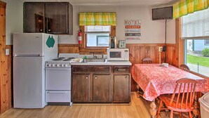 Une petite cuisine équipée d’un réfrigérateur, d’une cuisinière, d’un four à micro-ondes et d’un évier. On y trouve une table recouverte d’une nappe et une chaise. Une pancarte indique qu’il s’agit d’une chambre de motel offrant des commodités telles que la climatisation, un téléviseur et une piscine.