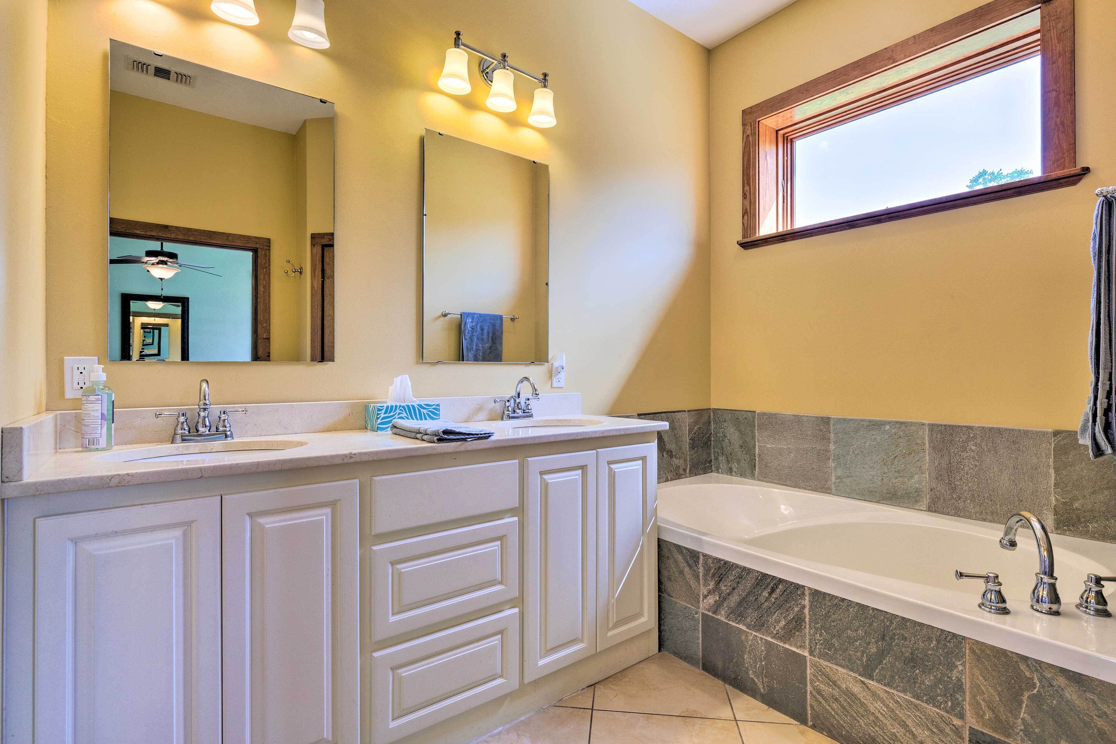 A bathroom with a double sink vanity, a large mirror, a bathtub, and a window.