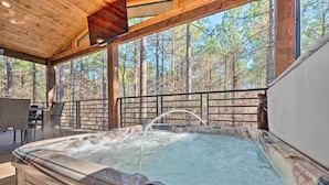 Un jacuzzi con caída de agua, rodeado de una terraza de madera y grandes ventanales con vistas a un bosque.