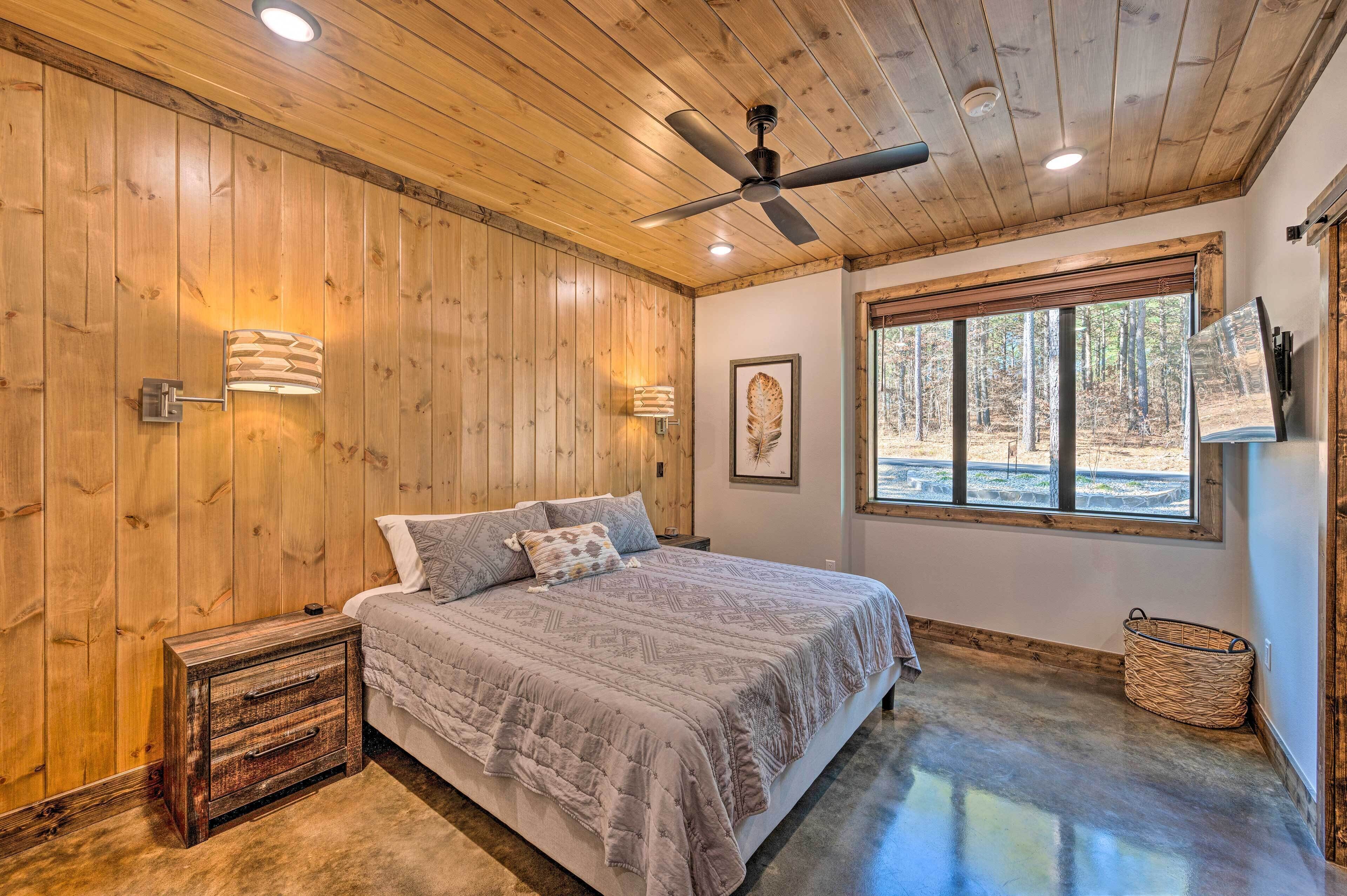 A bedroom with a wooden ceiling, a bed with a patterned bedspread, a ceiling fan, and a window with a view of trees.