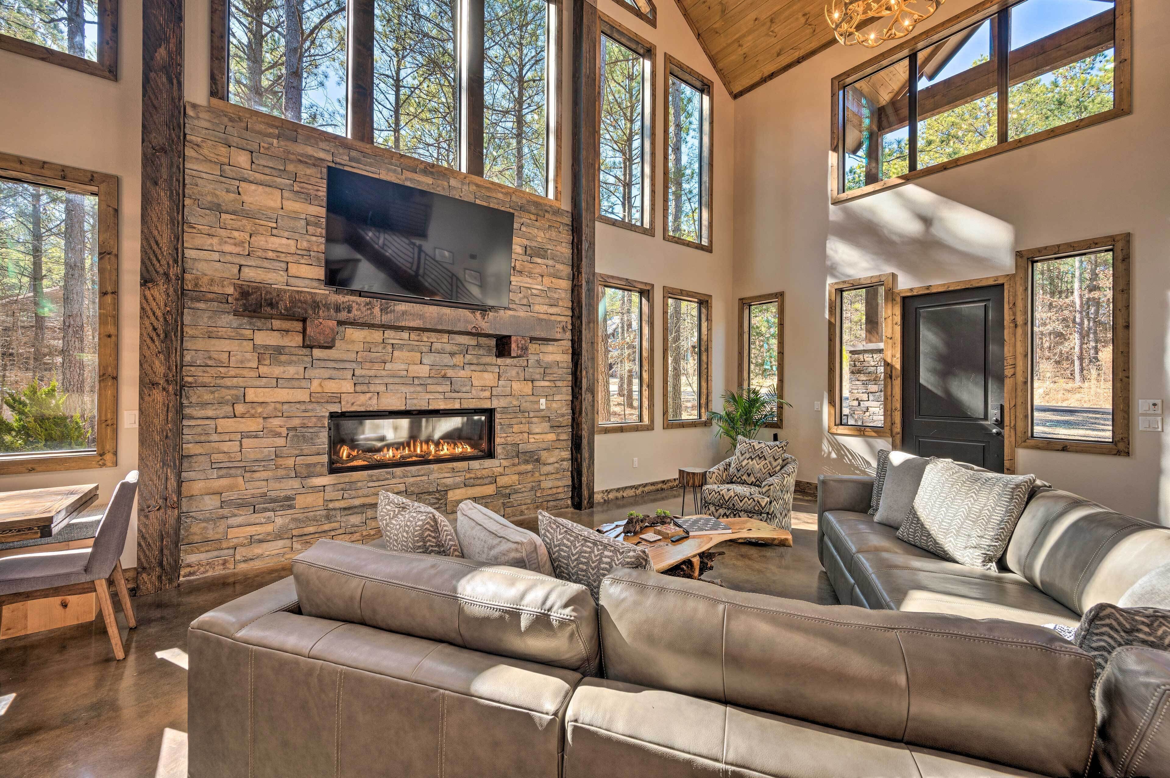 A spacious living room with a stone fireplace, leather sofas, and large windows offering views of the forest.