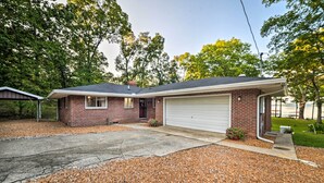 A single-story brick house with a garage, gravel driveway, and a small garden. - Charming Lakefront Home w/ Grill & Fire Pit! (New Concord)