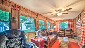 A rustic room with wooden walls, leather sofas, a ceiling fan, and large windows. - Charming Lakefront Home w/ Grill & Fire Pit! (New Concord)
