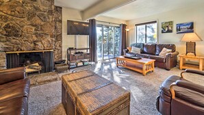 A cozy living room with a stone fireplace, leather sofas, a flat-screen TV, and a large window with a view of trees. - Slope Side Durango Condo at Purgatory Resort! (Durango)