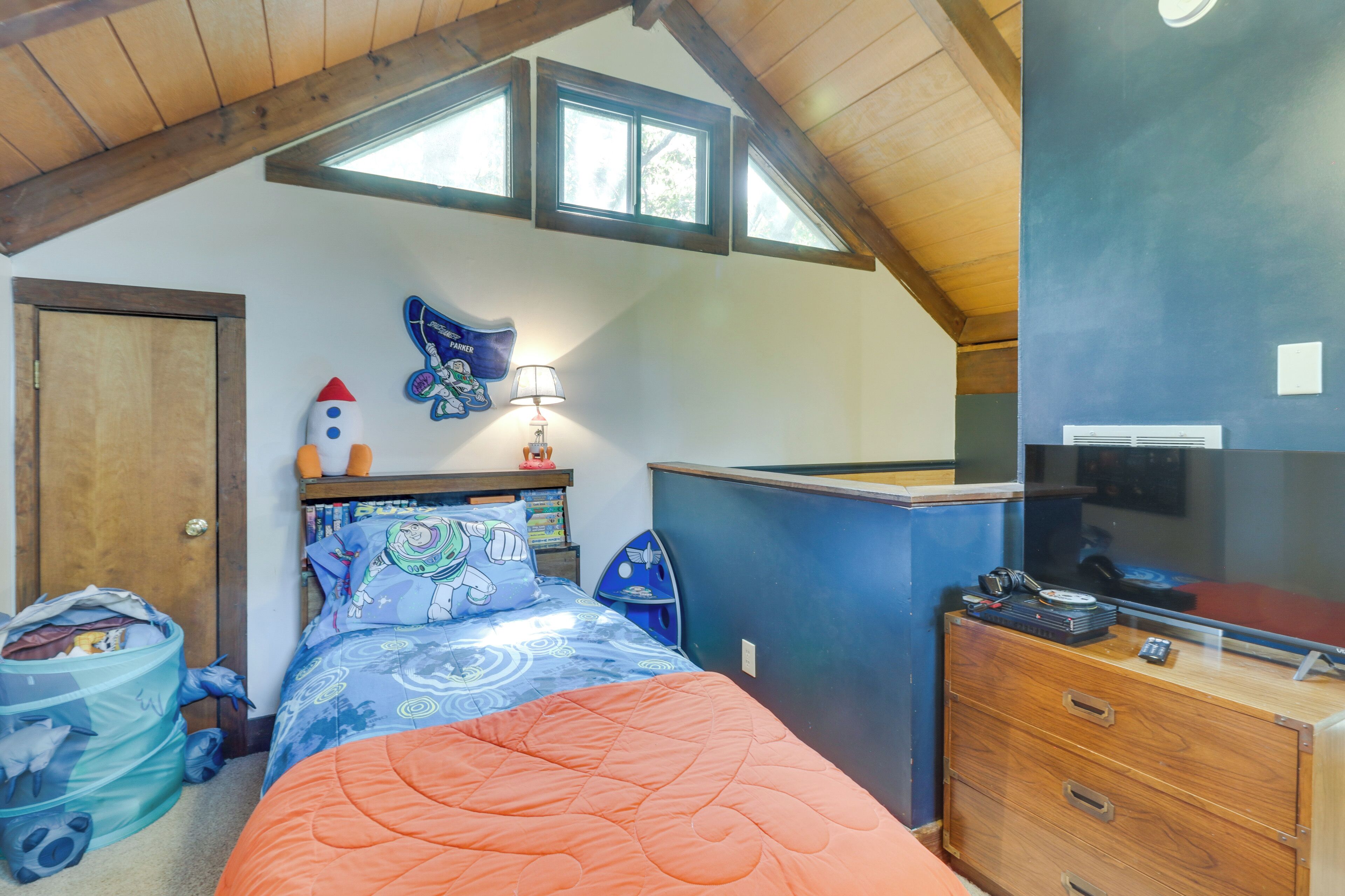 A bedroom with a sloped ceiling, a bed with a blue and orange bedspread, a small desk with a lamp, and a TV on a dresser.