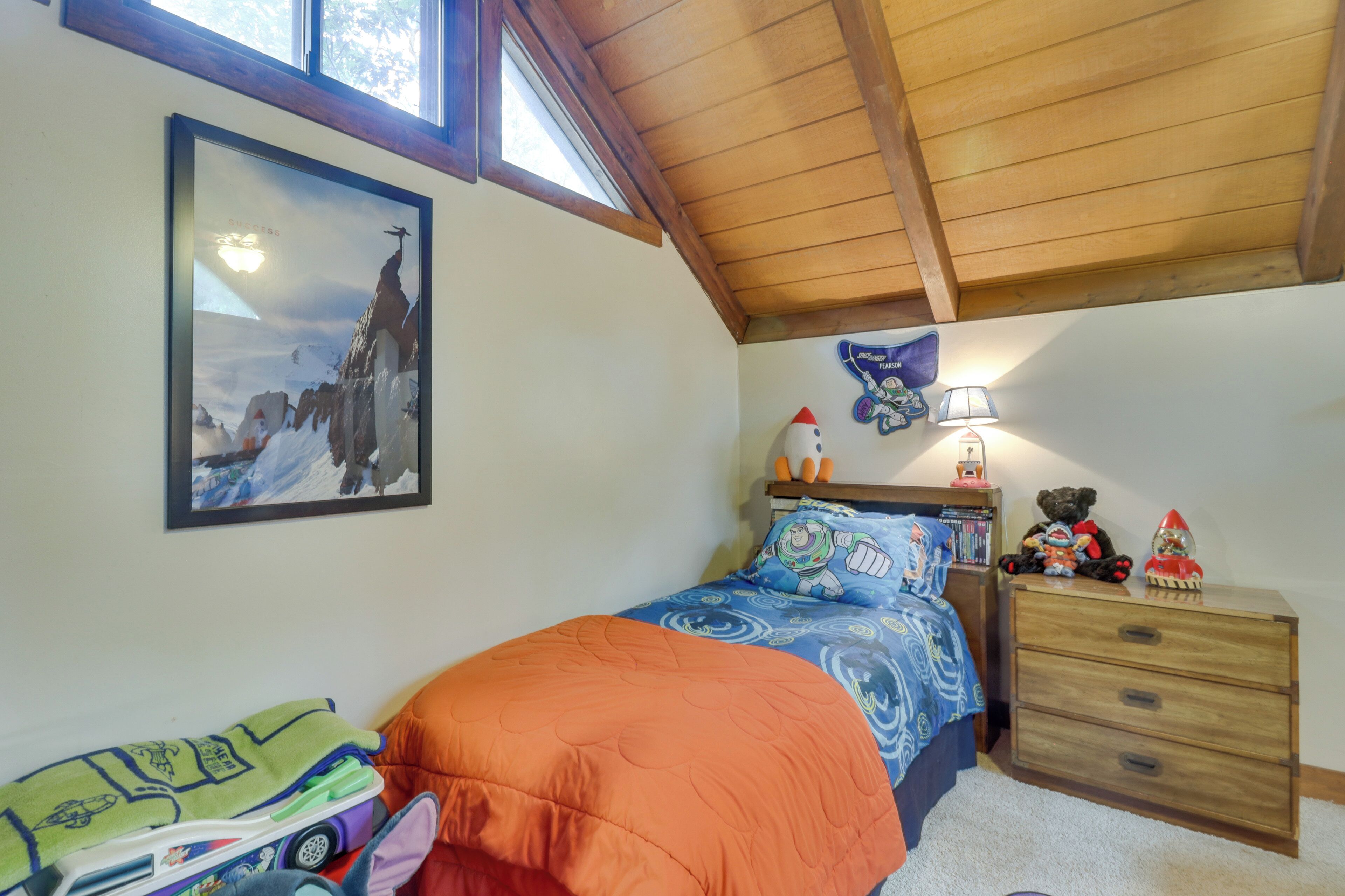Une chambre avec un plafond en bois, un lit recouvert d’une couverture orange, une table de chevet avec une lampe, un ours en peluche et un tableau encadré accroché au mur.