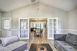 Un vaste salon doté d’un haut plafond, un coin repas avec une table et des chaises en bois, et une cuisine visible à travers une porte ouverte.