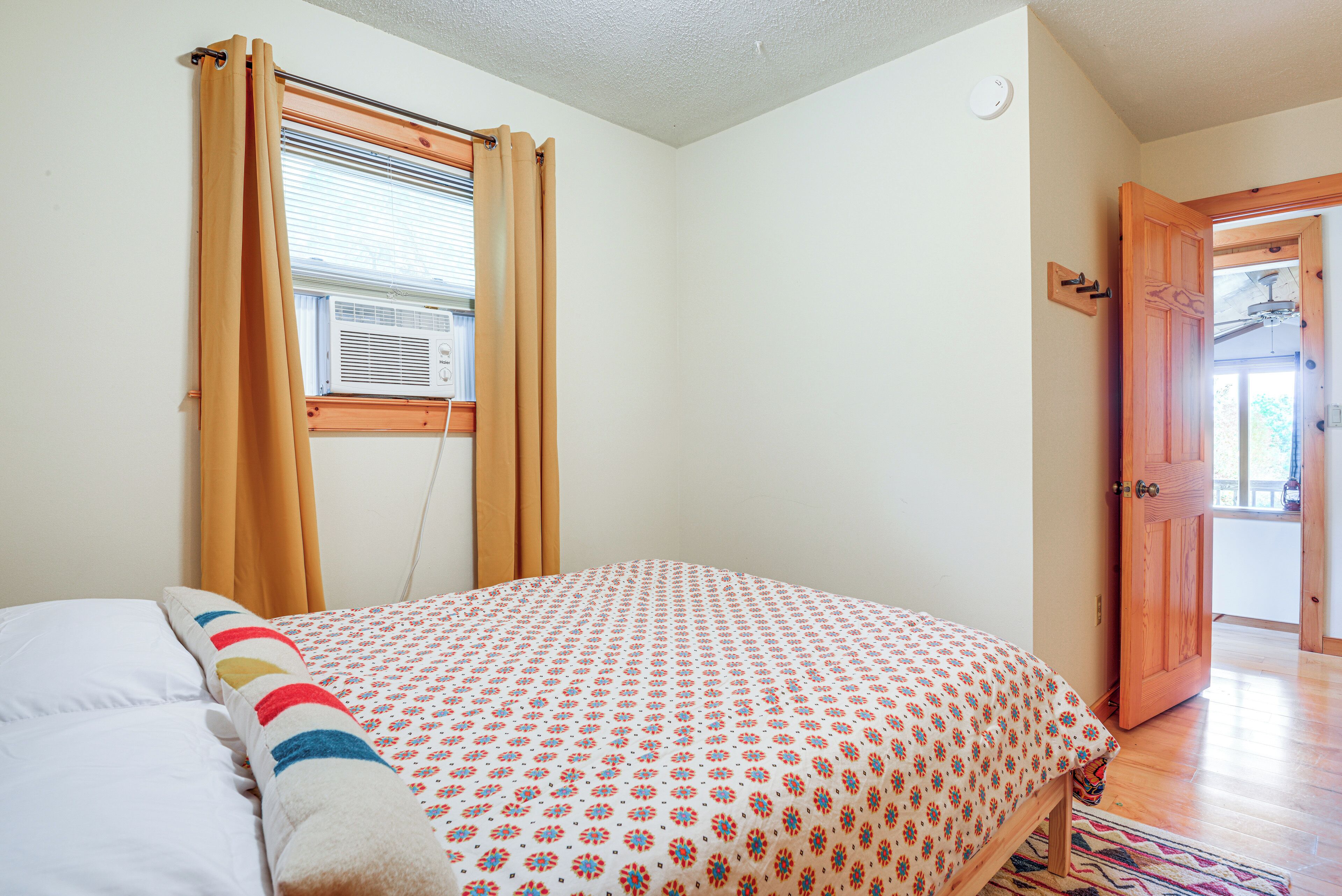 A bedroom with a bed, a window with curtains, an air conditioning unit, and a wooden door.