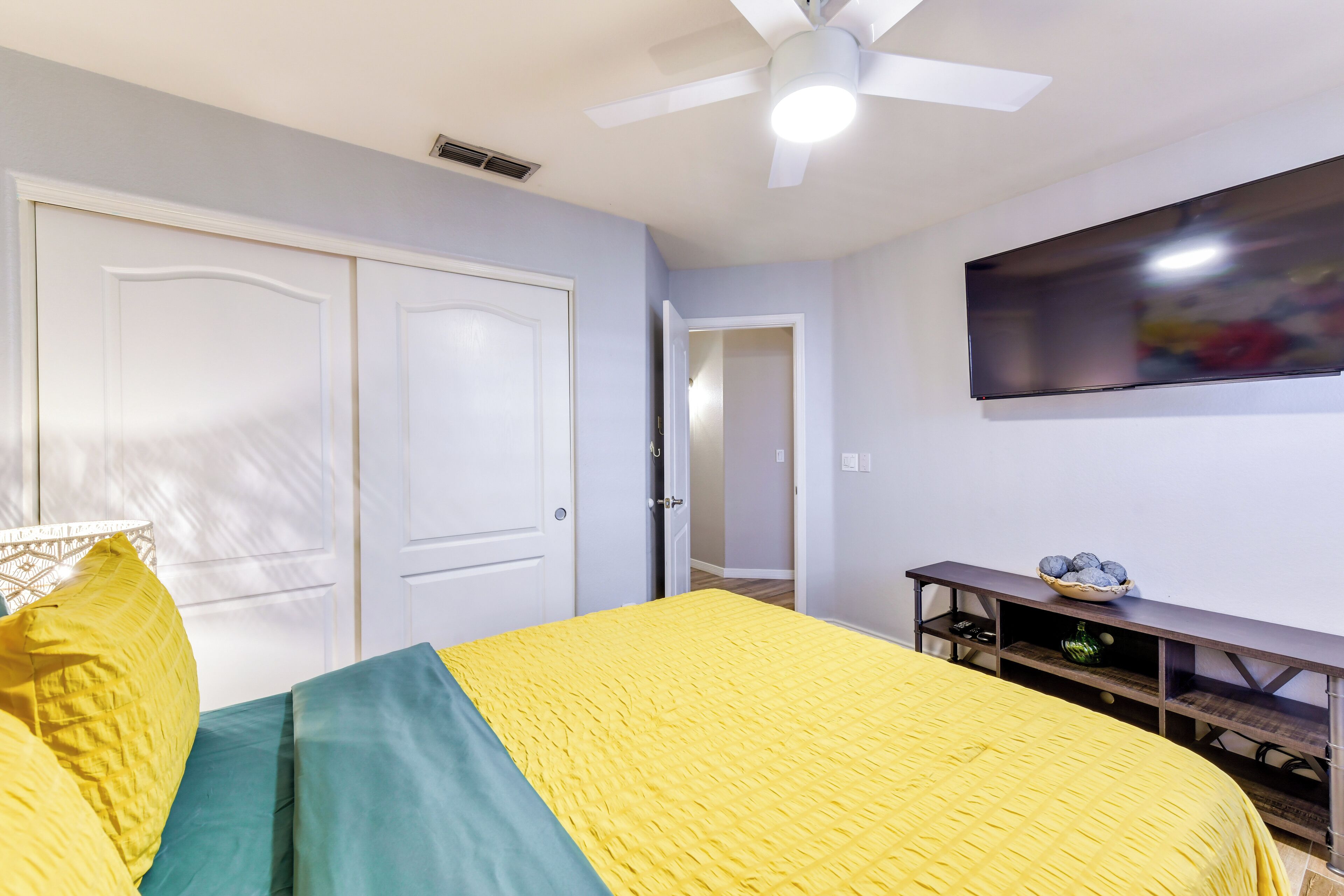 Une chambre à coucher avec un lit, un ventilateur de plafond, un téléviseur fixé au mur et une porte donnant sur une autre pièce.
