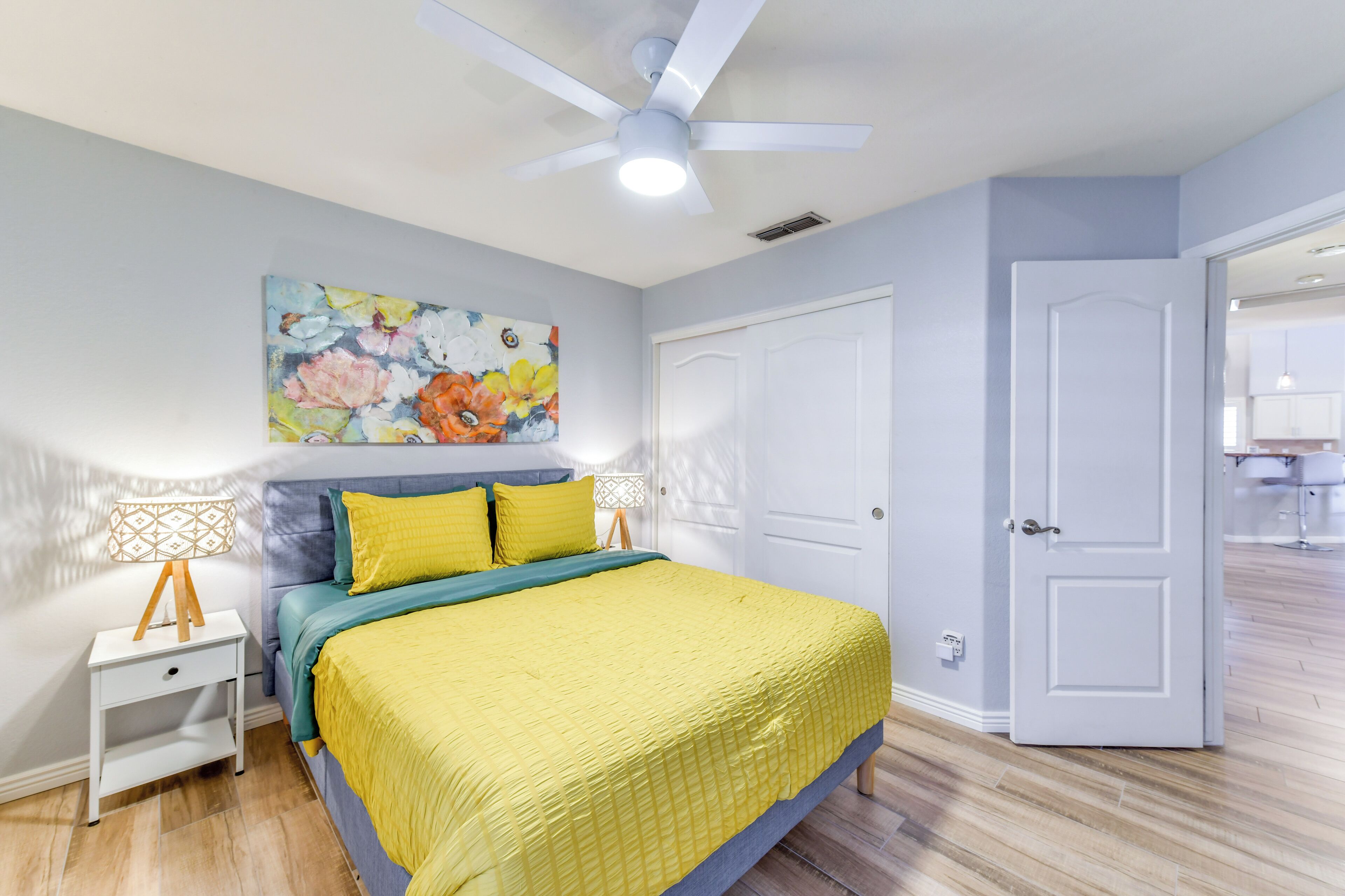 A bedroom with a bed, a ceiling fan, a painting, and a door leading to another room.
