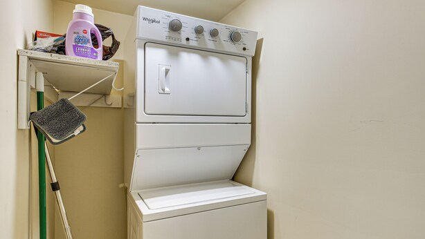 Une petite buanderie équipée d’un lave-linge et d’un sèche-linge superposés, d’une serpillière verte et d’une bouteille de lessive posée sur une étagère.
