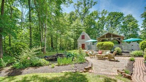 A rustic outdoor patio with stone flooring, surrounded by lush greenery and a wooden fence. - Cozy Virginia Vacation Rental w/ Seasonal Pool! (Nokesville)