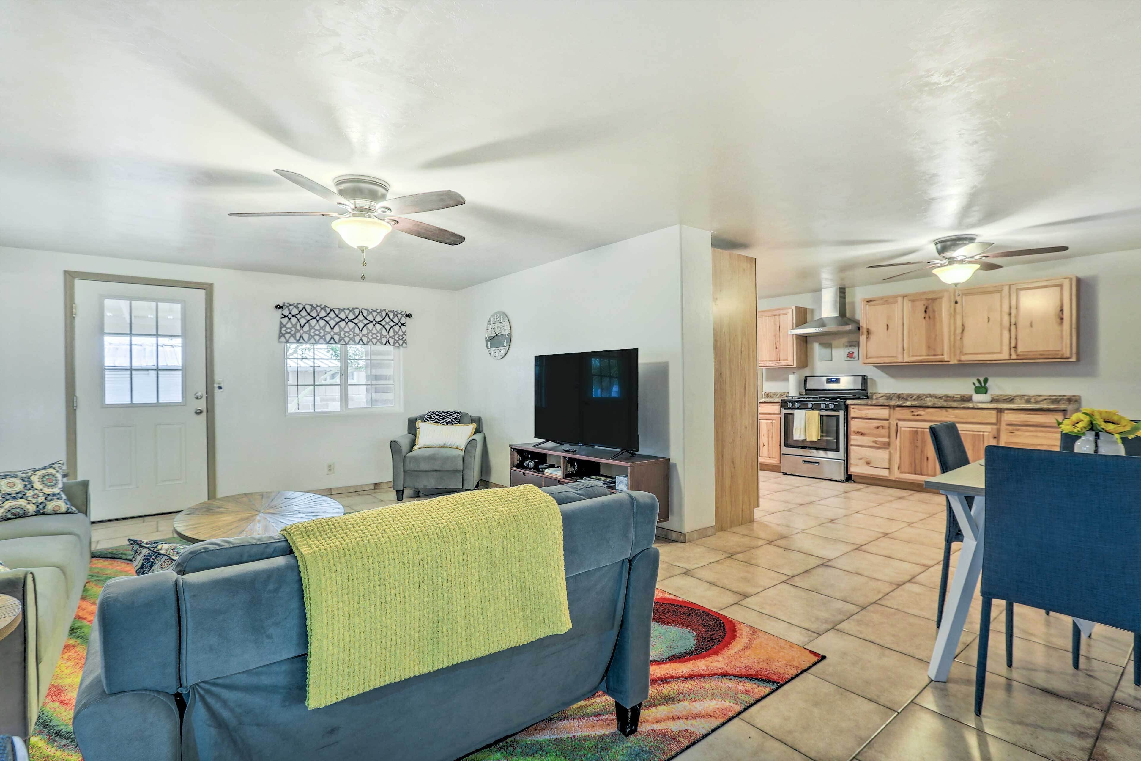 A living room with a sofa, TV, and ceiling fan. There is a kitchen area with cabinets and a dining table.