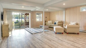 A spacious living room with a wooden floor, a beige sofa set, and a large sliding glass door leading to an outdoor area. - Lakefront Cabin w/ Panoramic Water View & Access (Sunapee)