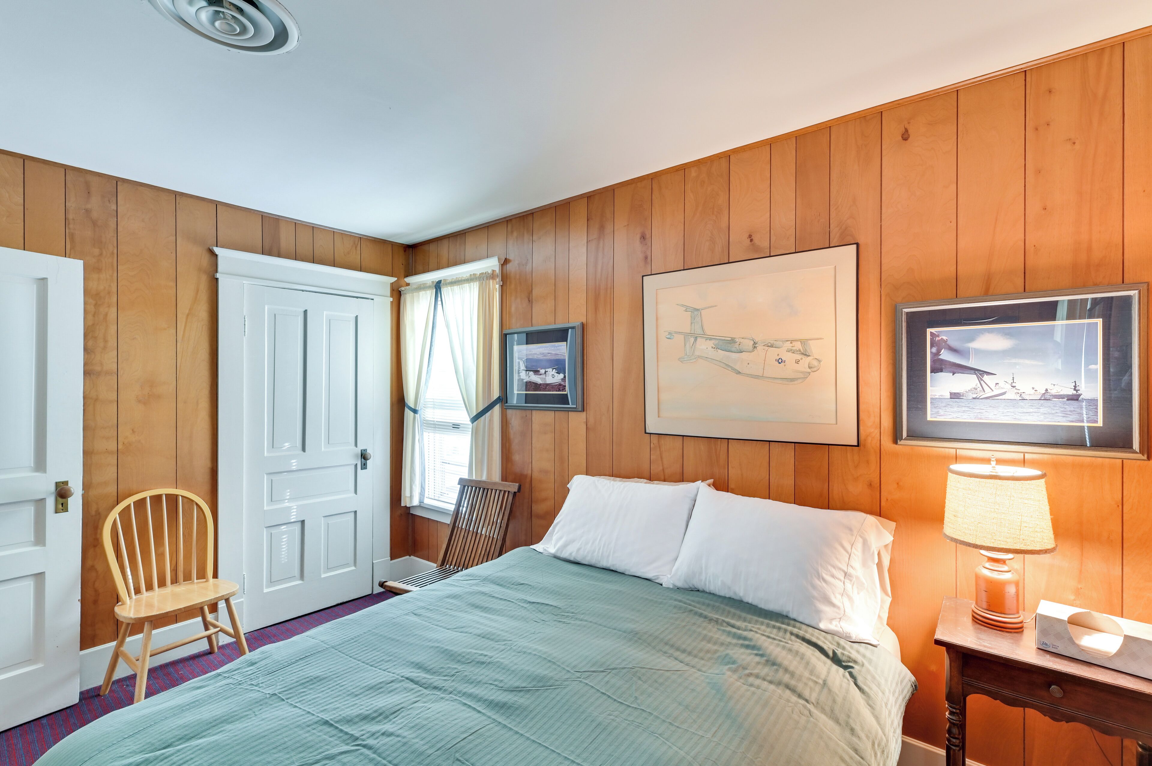 A room with wooden paneling, a bed with white linens, a chair, a table with a lamp, and two framed pictures on the wall.