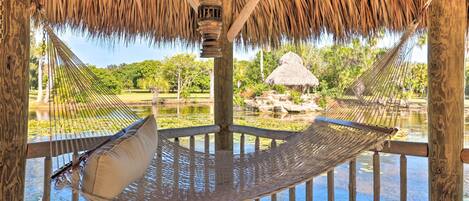 A hammock under a thatched roof structure with a view of a water body and greenery.