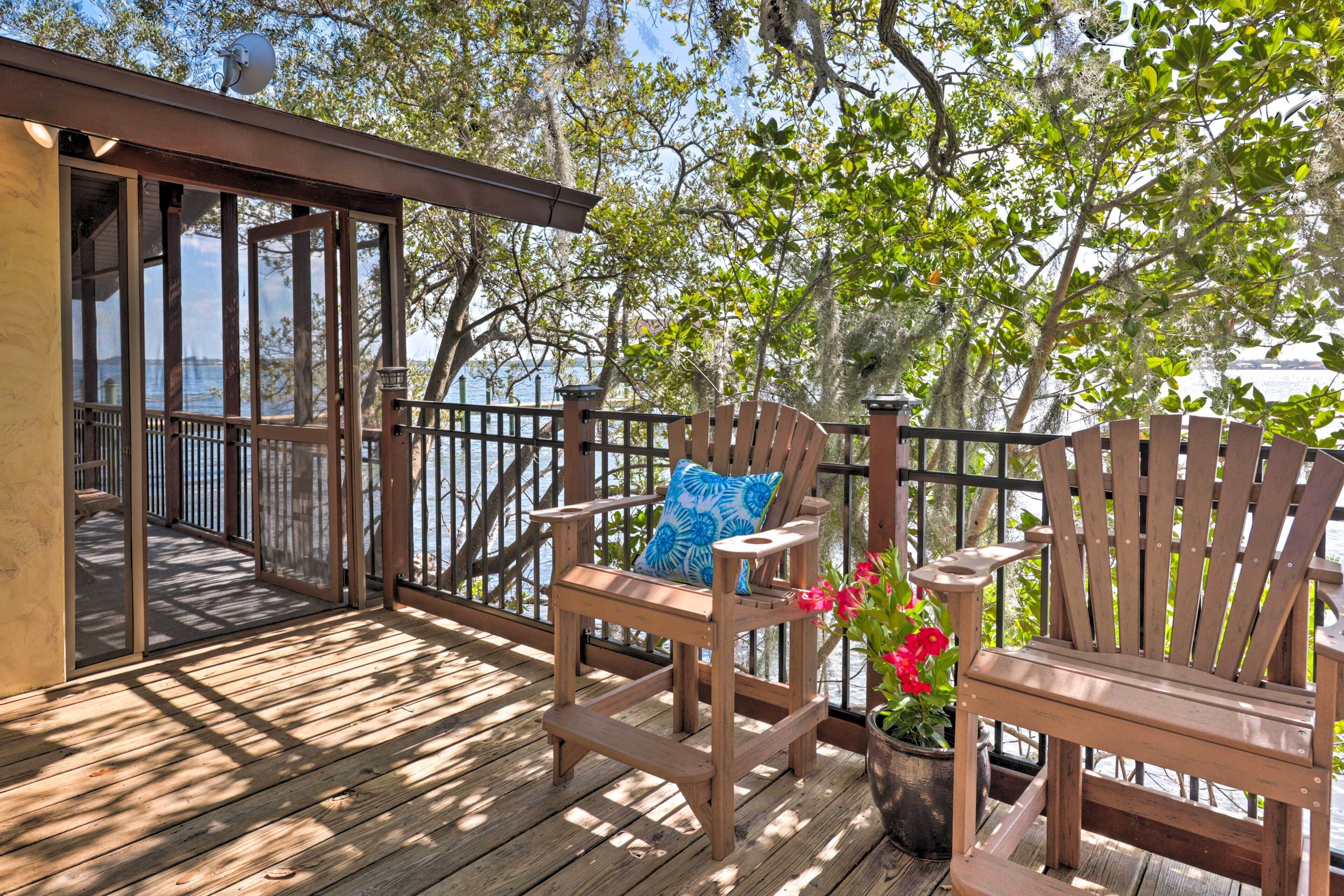 A wooden deck with two Adirondack chairs, one with a pillow, and a potted plant with red flowers.
