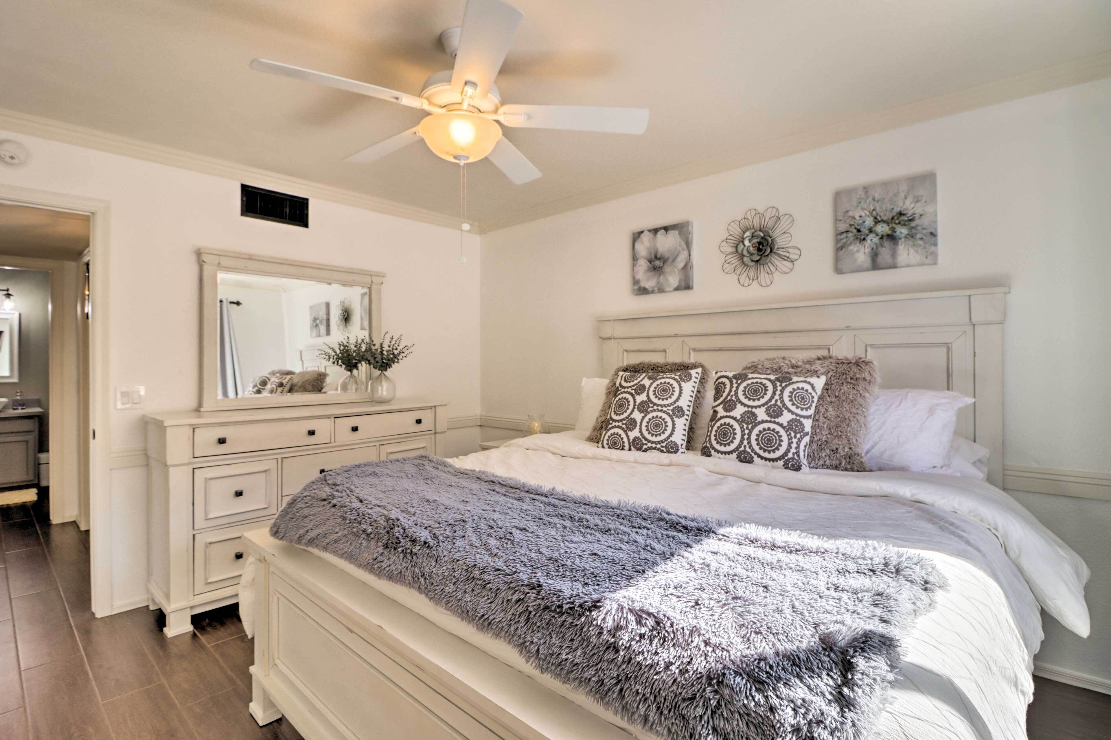 Une chambre à coucher comprenant un lit, une commode, un miroir, un ventilateur de plafond et des œuvres d’art murales.