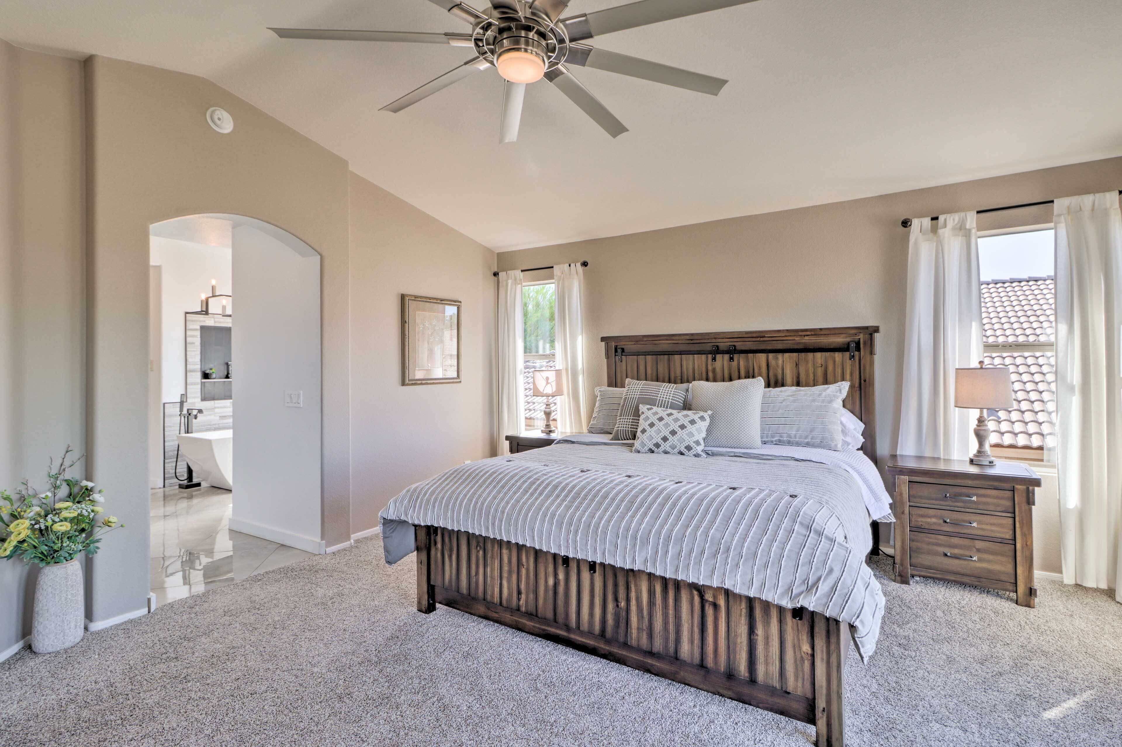 A bedroom with a large bed, a ceiling fan, a nightstand, and a view of the outside.