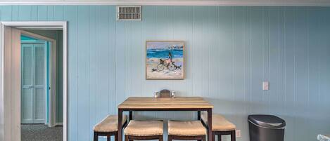 A dining area with a wooden table and four chairs, a painting on the wall, a trash can, and a door leading to another room.