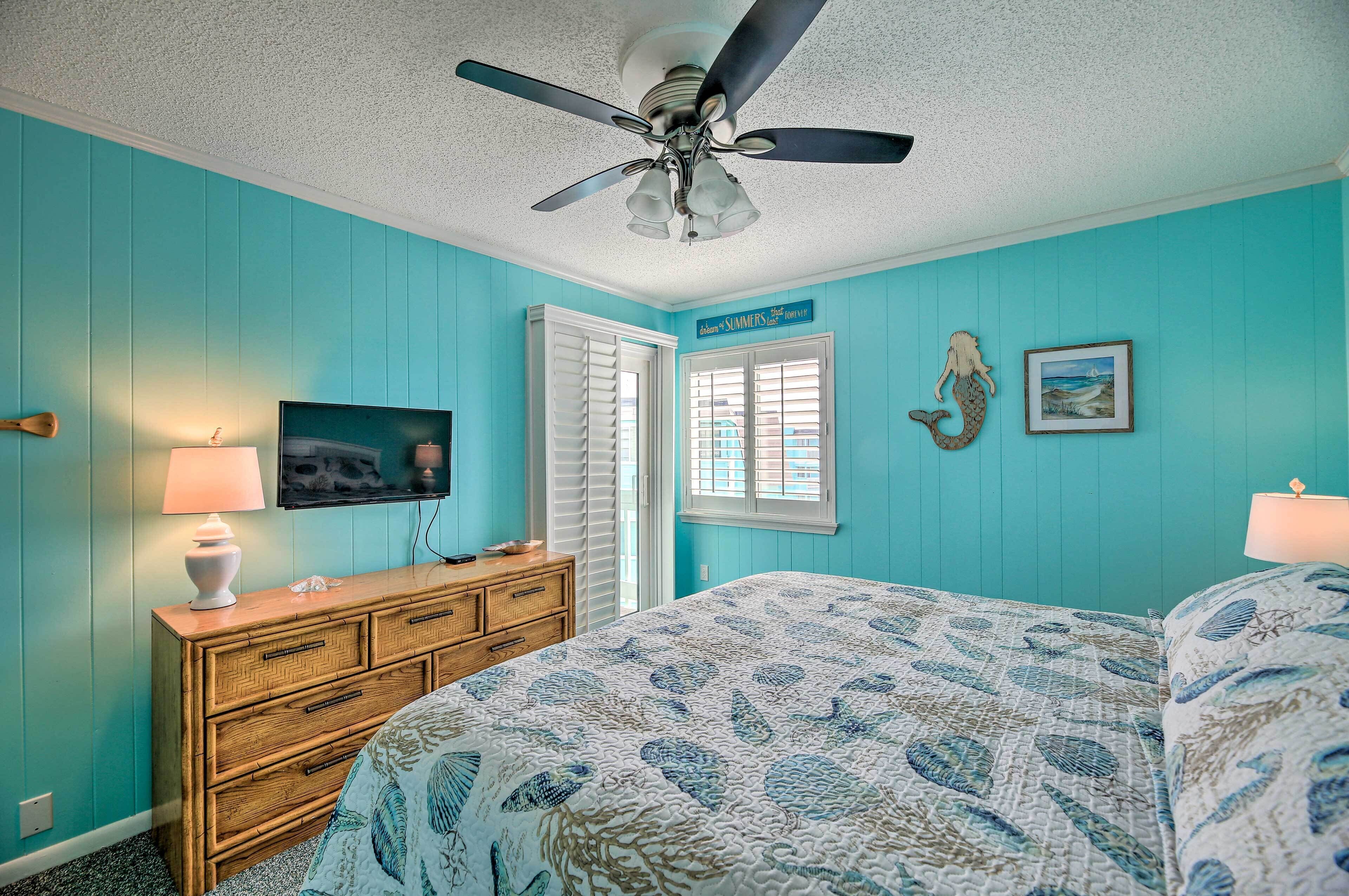 A bedroom with teal walls, a bed with a blue and white patterned bedspread, a wooden dresser, a ceiling fan, a television, and a window with shutters.