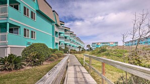 A wooden boardwalk leading to a row of turquoise buildings with balconies, surrounded by greenery. - Beachfront Condo w/ Boardwalk & Pool Access! (Atlantic Beach)