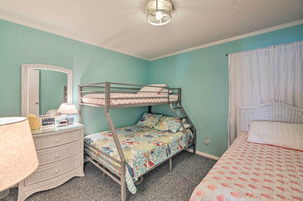 A bunk bed with a colorful bedspread, a dresser with a lamp, a mirror, and a window with curtains.