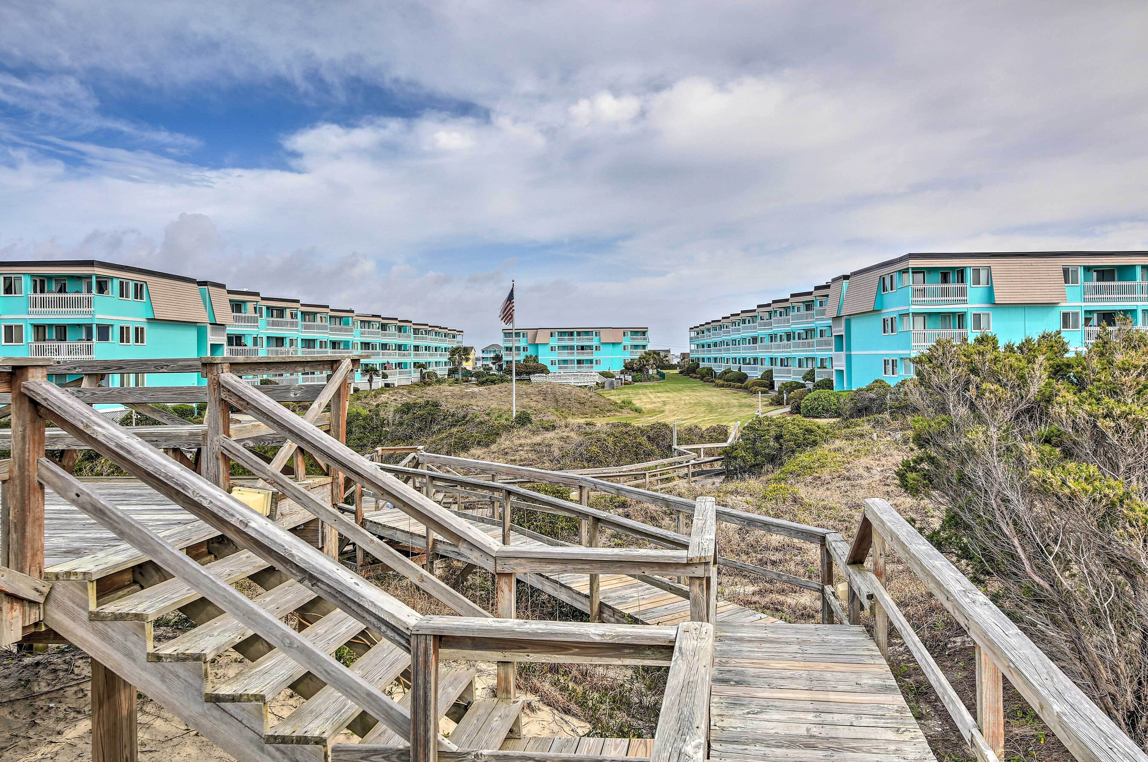 A wooden boardwalk leading to turquoise buildings with a flag, surrounded by natural landscape.