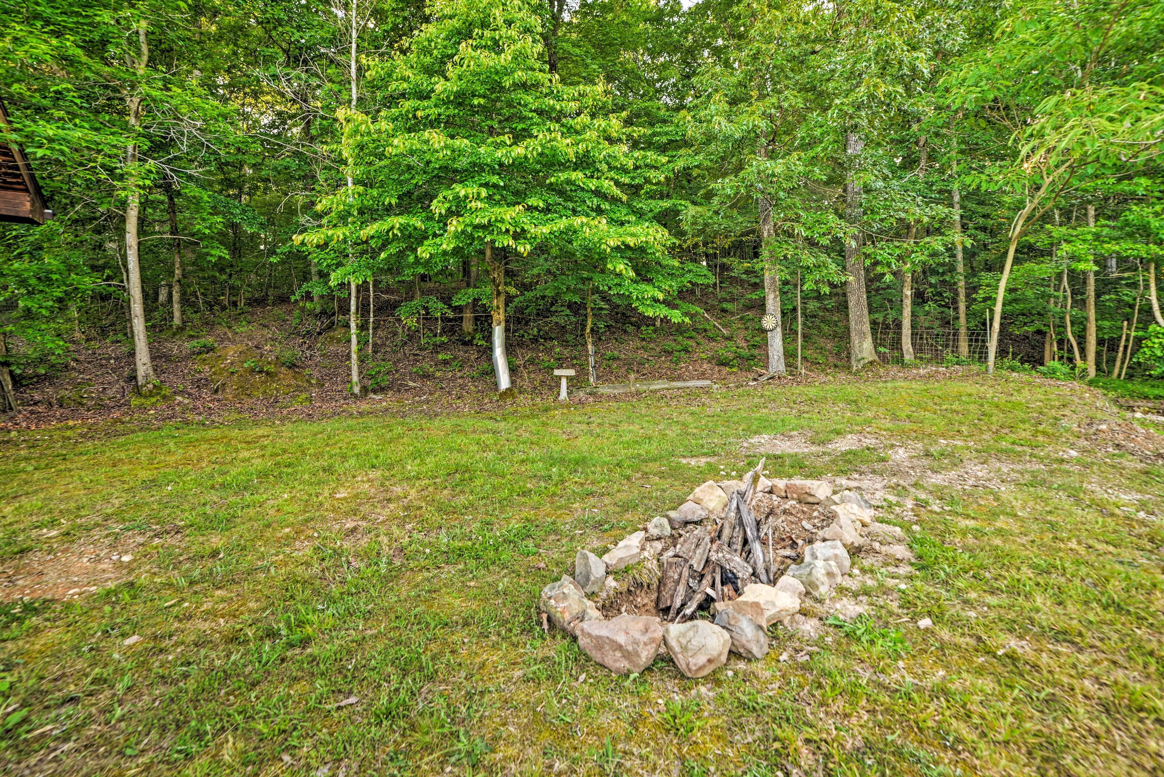 A forested area with a fire pit surrounded by rocks and logs, a wooden structure in the background, and a fenced area.