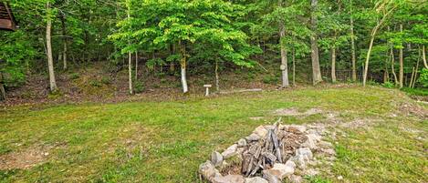 A forested area with a fire pit surrounded by rocks and logs, a wooden structure in the background, and a fenced area.