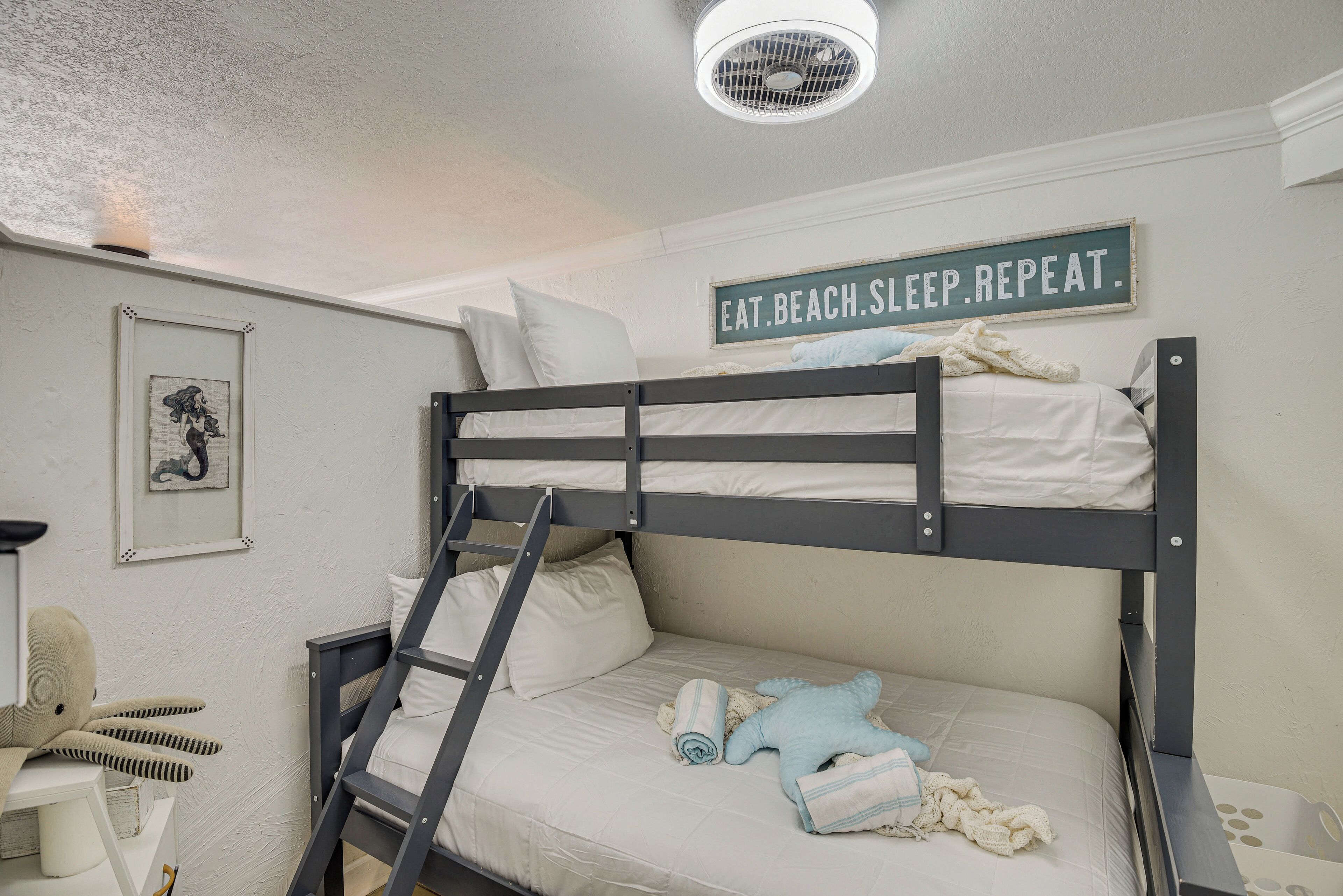 Un lit superposé avec une échelle, un tableau encadré et une pancarte sur laquelle on peut lire : « MANGER. SE PRÉLASSER À LA PLAGE. DORMIR. RECOMMENCER. »