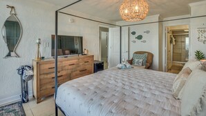 A bedroom with a large bed, a wooden dresser, a TV, and a chandelier. - Walk to Shore! Top-floor Daytona Beach Condo (Daytona Beach Shores)