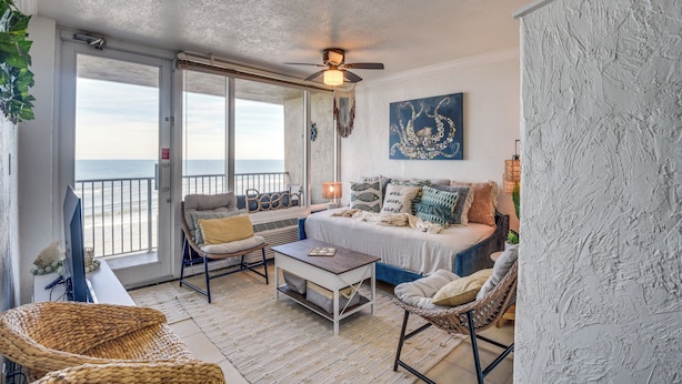 A beachside room with a balcony, a ceiling fan, a bed with multiple pillows, a coffee table, and a wicker chair. - Walk to Shore! Top-floor Daytona Beach Condo (Daytona Beach Shores)