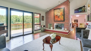 A spacious living room with a fireplace, a large painting, and a balcony with a view of trees. - Pet-friendly Vacation Rental w/ Lake Access (Newton)