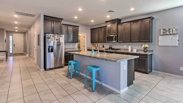 A modern kitchen with dark cabinets, a stainless steel refrigerator, a microwave, and a granite island with two blue bar stools. - Quiet Family Home w/ Yard ~ 3 Mi to Luke Afb! (Glendale)