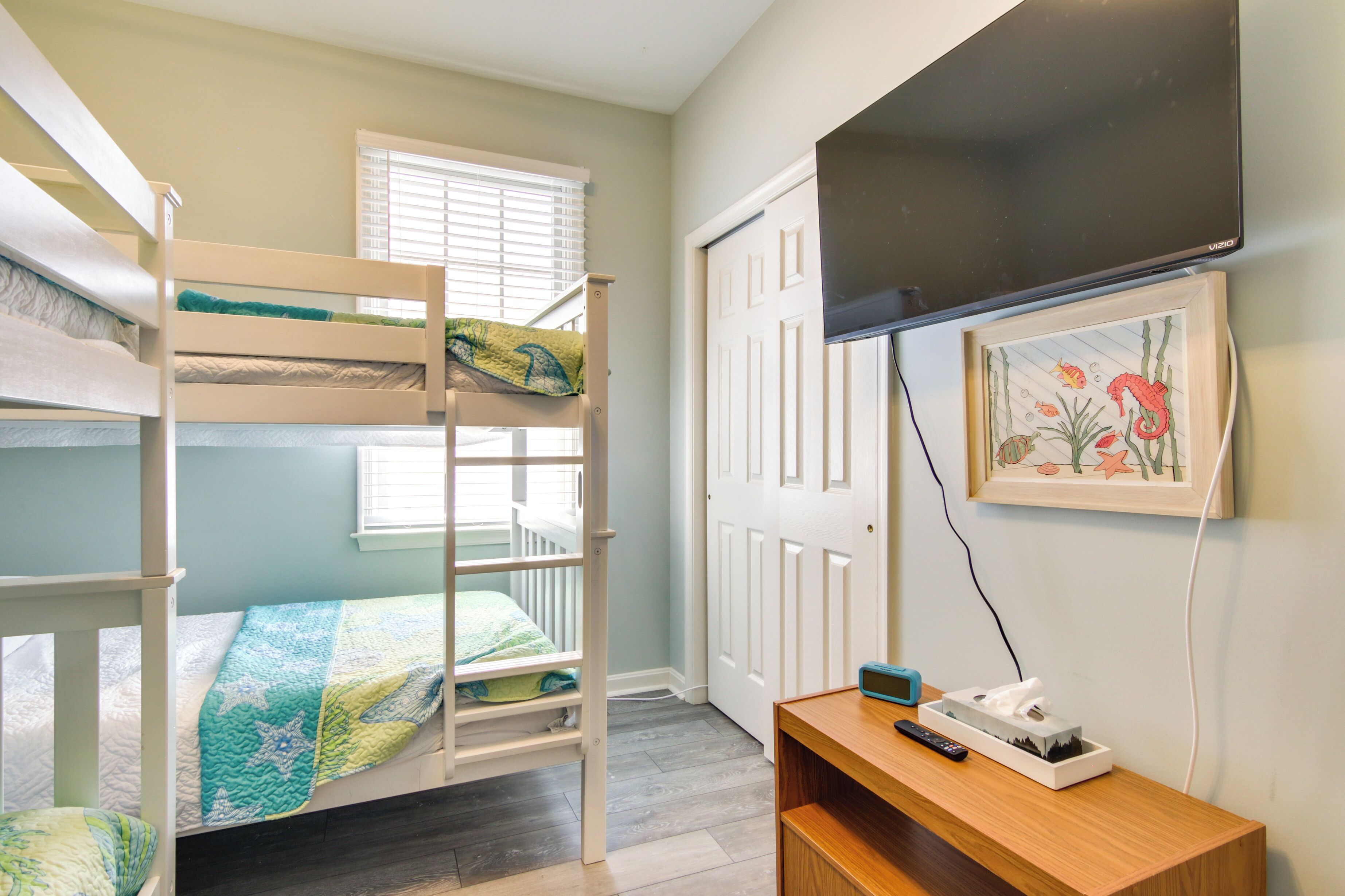 A bunk bed room with a TV mounted on the wall, a framed picture, and a bedside table with a remote control and tissues.