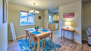 A cozy living room with a wooden dining table, blue placemats, wicker chairs, a chandelier, and a sign that reads "Welcome to the COTTAGE." - Anacortes Condo - Walk to Town, Marina, + Bay! (Anacortes)
