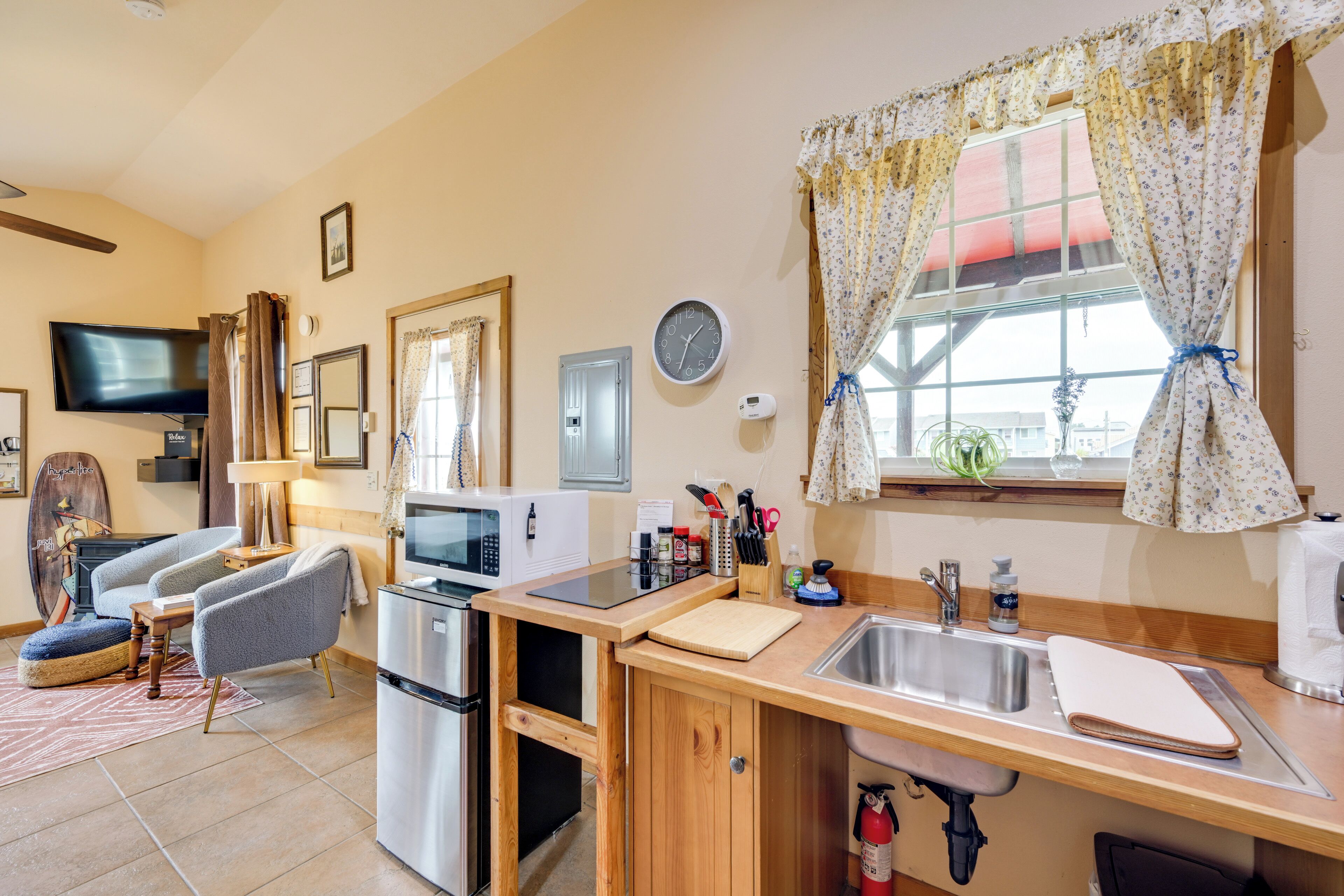 A compact kitchen with a microwave, refrigerator, and sink. There is a television mounted on the wall, and a window with curtains.
