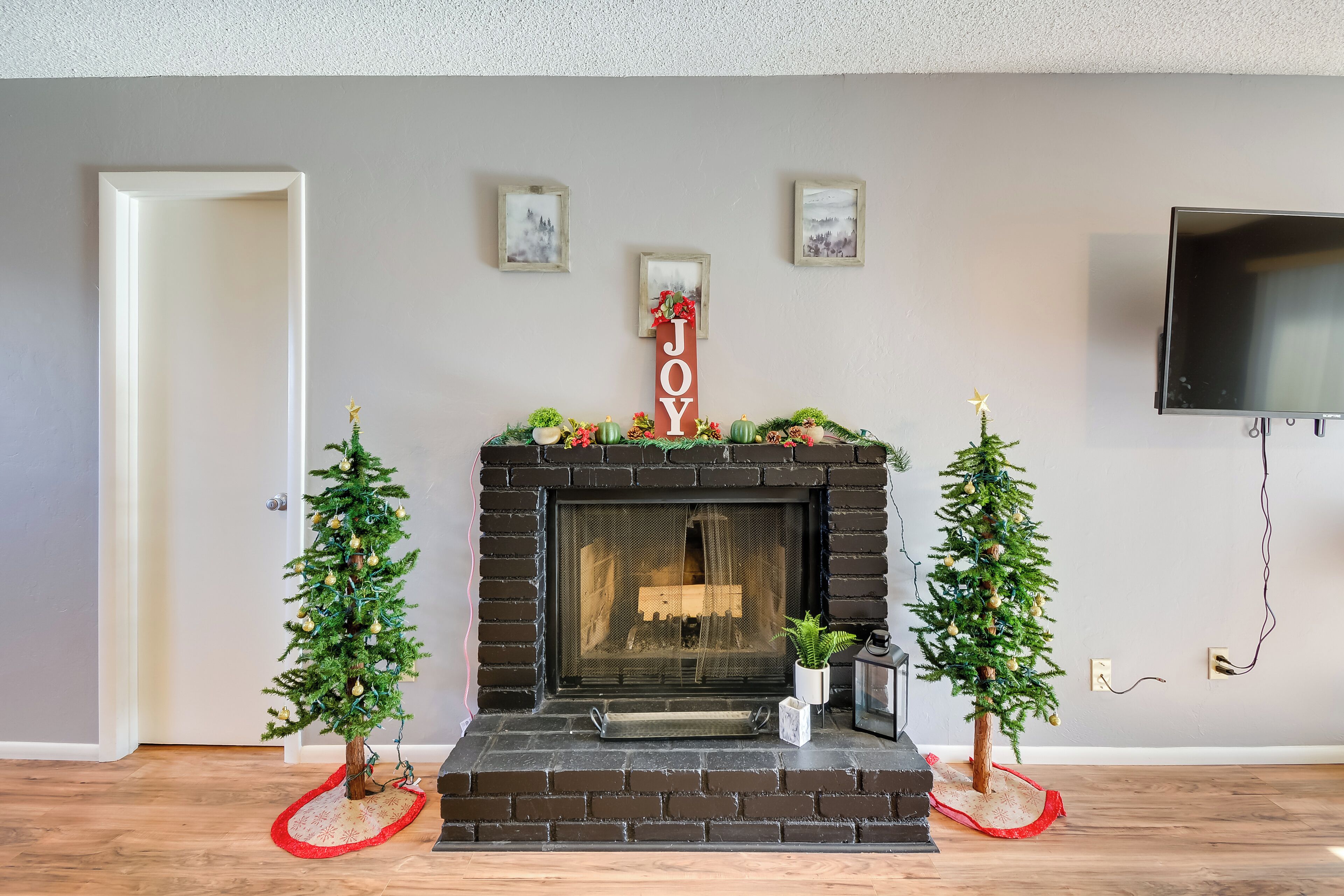 Una chimenea con un letrero que dice 'JOY' encima, flanqueada por dos pequeños árboles de Navidad, y un televisor montado en la pared.