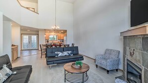 Ein geräumiges Wohnzimmer mit einer schwarzen Ledersofa, einem gemusterten Sessel und einem runden Couchtisch mit einer Pflanze. Im rechten Bildbereich ist ein Kamin zu sehen, und im Hintergrund befindet sich eine Küchenzeile.
