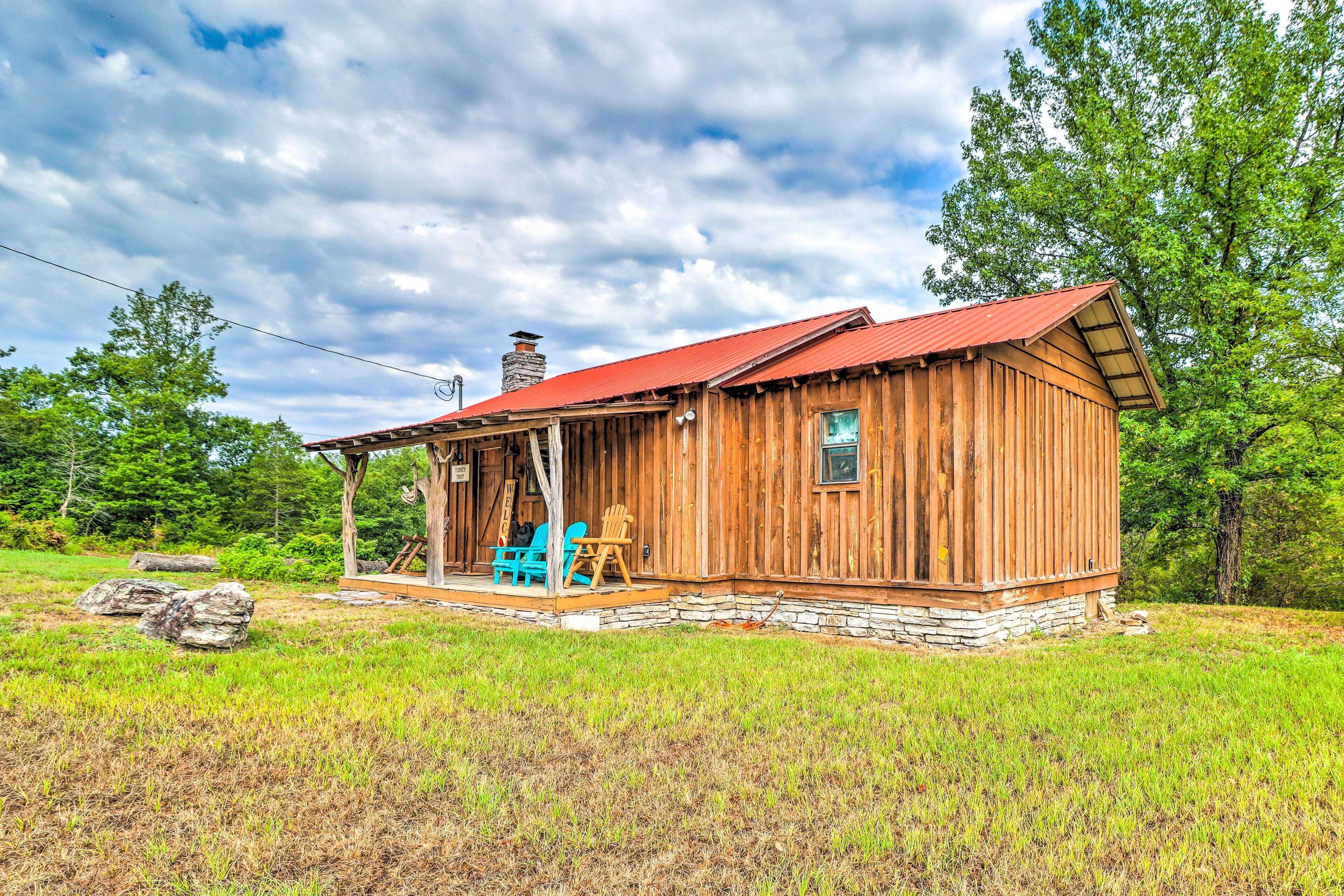 A wooden cabin with a red roof, a porch with blue chairs, and a stone foundation.