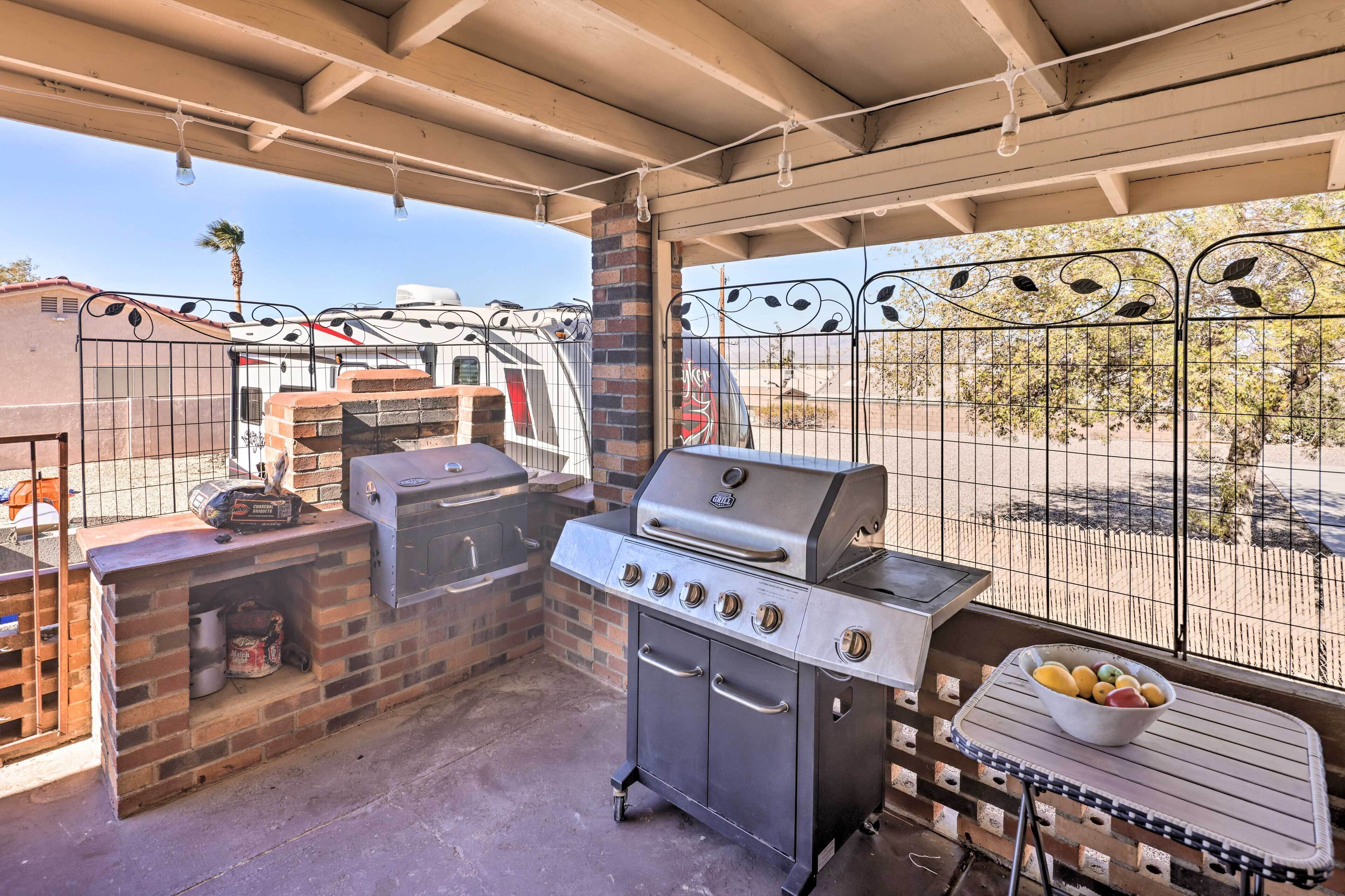 A covered outdoor grilling area with a brick barbecue, a table with a bowl of fruit, and a view of a fenced yard.