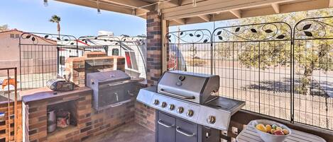 A covered outdoor grilling area with a brick barbecue, a table with a bowl of fruit, and a view of a fenced yard.