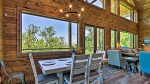 A wooden cabin interior with a dining table, chairs, a sofa, and a large window offering a view of trees.