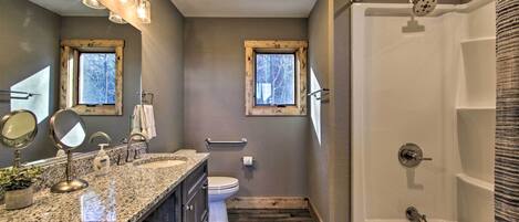 A bathroom with a granite countertop, two sinks, a toilet, a shower, and two windows.
