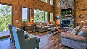 A spacious log cabin interior with a stone fireplace, multiple seating areas, and large windows offering a view of the surrounding forest.