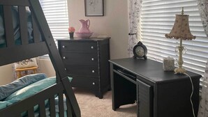 A bedroom with a wooden bunk bed, a dresser with drawers, a desk with a lamp, a clock, and a framed picture on the wall.